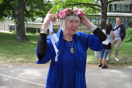 Birgitta Stenberg har en kornblå lång sommarjacka och ett stort halssmycke. Håret, som hon klipper själv, är i en kort page med lugg.Hon rättar till blomsterkransen på huvudet.